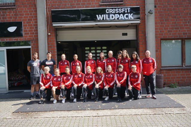 Gruppe von 15 Sportlern in roten Trainingsanz&uuml;gen vor dem Eingang von CrossFit Wildpack, umgeben von Ziegelbauten.