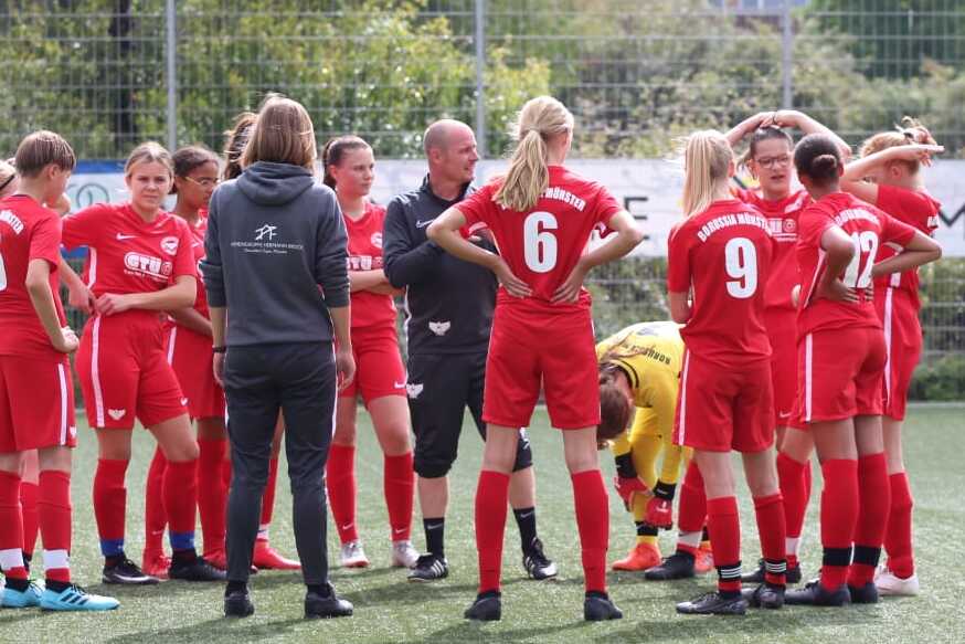 Frauenfu&szlig;ballmannschaft in roten Trikots versammelt sich um den Trainer auf einem Kunstrasenplatz.