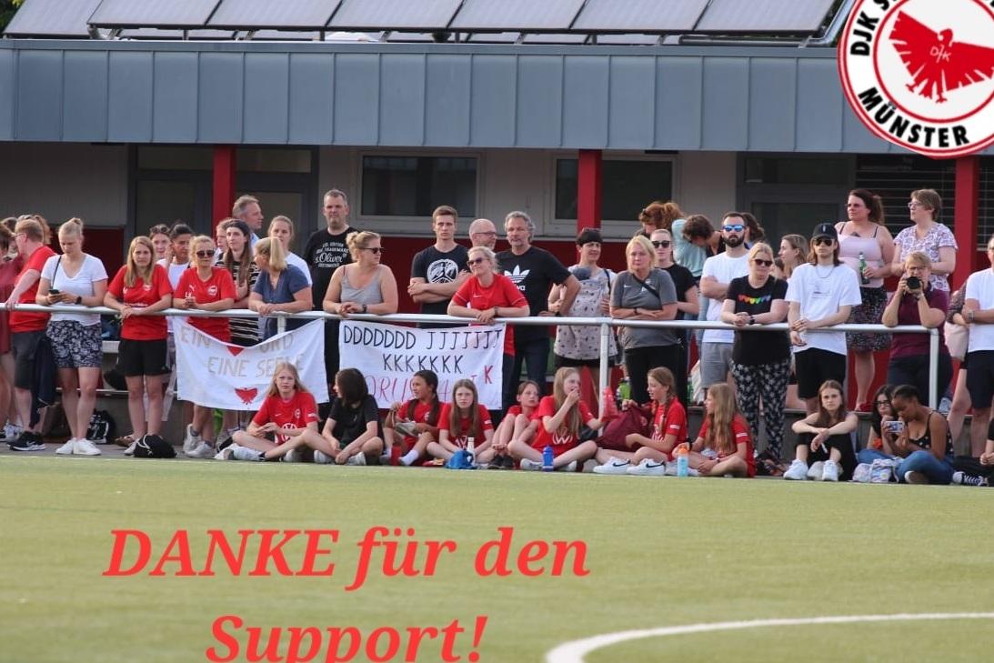 Eine Gruppe von Zuschauern mit roten Trikots steht an einem Fu&szlig;ballfeld, einige halten Banner mit Dankesschriften.