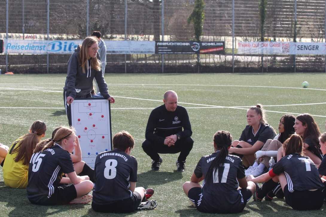 Trainer und Spielerinnen sitzen im Kreis auf dem Fu&szlig;ballplatz; die Trainerin zeigt Taktik auf einem wei&szlig;en Board.