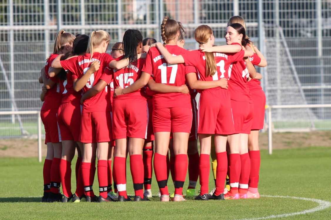 Gruppe von Fu&szlig;ballspielerinnen in roten Trikots umarmt sich auf dem Spielfeld vor einem Spiel.