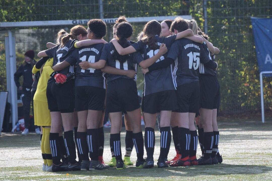 Gruppe von Fu&szlig;ballspielerinnen steht umarmt zusammen auf einem Sportplatz, bereit f&uuml;r das Spiel.