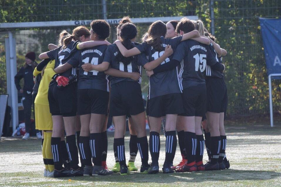Gruppe von Fu&szlig;ballspielerinnen steht umarmt zusammen auf einem Sportplatz, bereit f&uuml;r das Spiel.