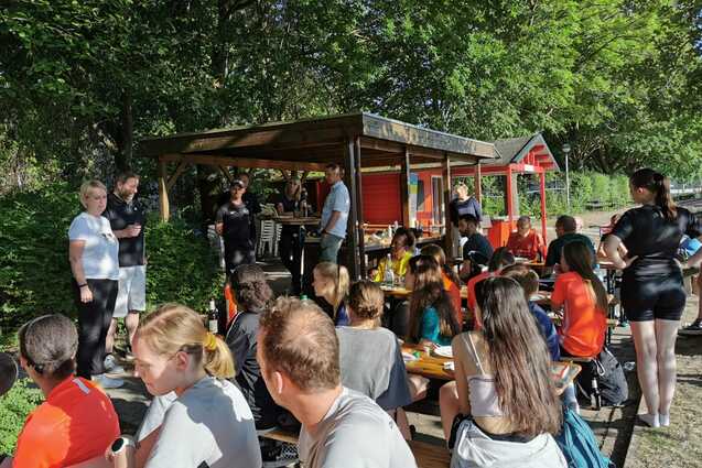 Gruppenszene mit Sportlern, die an Tischen sitzen und essen, umgeben von Gr&uuml;n und einem gem&uuml;tlichen Kiosk.