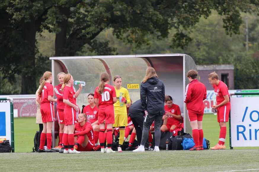 Mannschaft sitzt auf der Ersatzbank, w&auml;hrend die Trainerin Anweisungen gibt; Spieler in roten Trikots auf einem Fu&szlig;ballplatz.