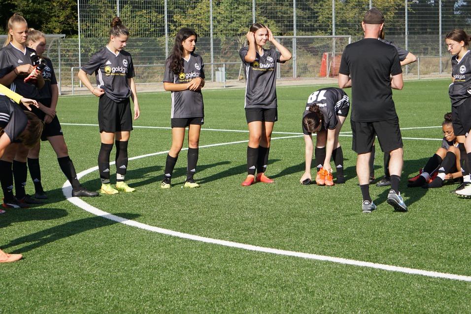 Mehrere Fu&szlig;ballspielerinnen in grauen Trikots versammeln sich um einen Trainer auf einem Rasenplatz.
