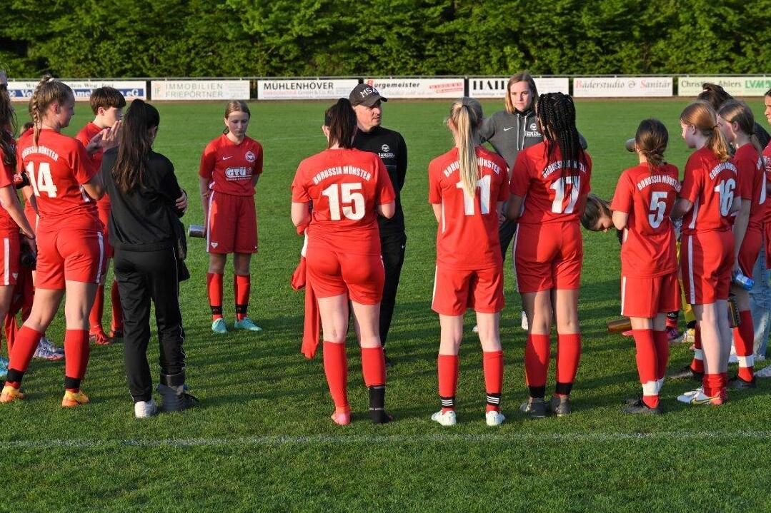 Gruppe von Fu&szlig;ballerinnen in roten Trikots versammelt sich um einen Trainer auf einem gr&uuml;nen Spielfeld.