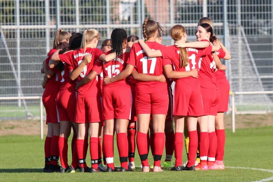 Gruppe von Fu&szlig;ballspielerinnen in roten Trikots umarmt sich auf dem Spielfeld vor einem Spiel.