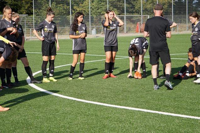 Mehrere Fu&szlig;ballspielerinnen in grauen Trikots versammeln sich um einen Trainer auf einem Rasenplatz.