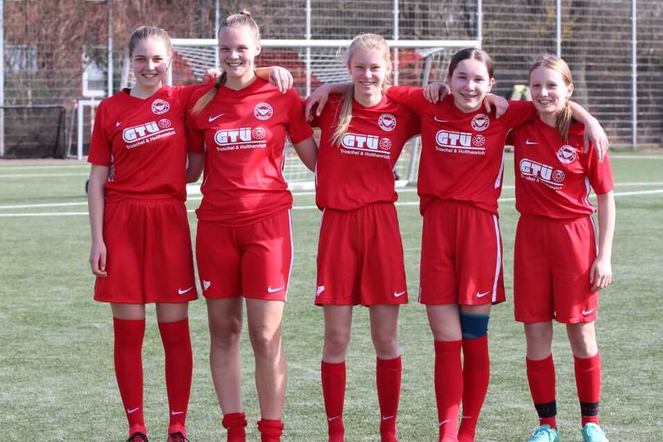 F&uuml;nf M&auml;dchen in roten Fu&szlig;balltrikots posieren gemeinsam auf einem Fu&szlig;ballfeld.