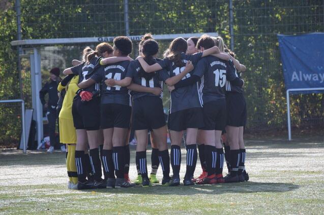 Gruppe von Fu&szlig;ballspielerinnen steht umarmt zusammen auf einem Sportplatz, bereit f&uuml;r das Spiel.
