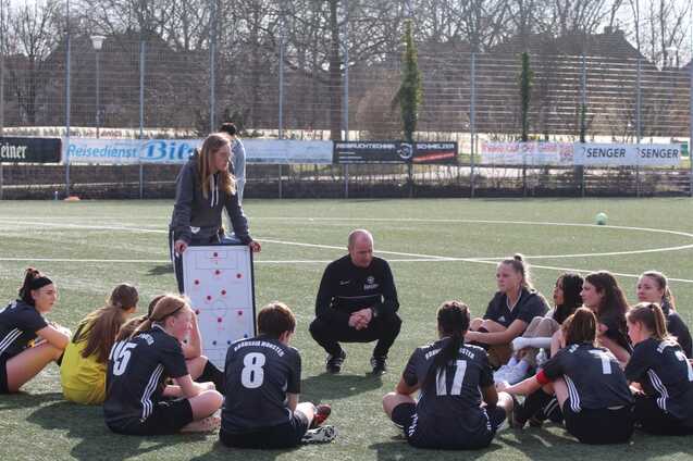 Trainer und Spielerinnen sitzen im Kreis auf dem Fu&szlig;ballplatz; die Trainerin zeigt Taktik auf einem wei&szlig;en Board.
