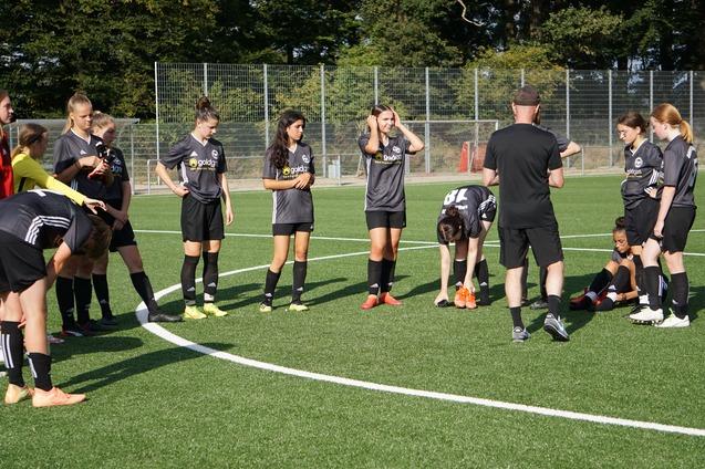 Mehrere Fu&szlig;ballspielerinnen in grauen Trikots versammeln sich um einen Trainer auf einem Rasenplatz.