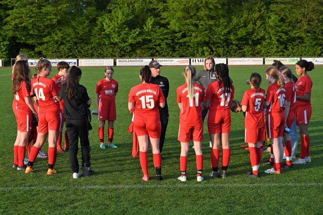 Gruppe von Fu&szlig;ballerinnen in roten Trikots versammelt sich um einen Trainer auf einem gr&uuml;nen Spielfeld.