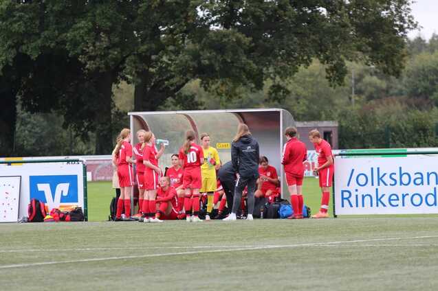 Mannschaft sitzt auf der Ersatzbank, w&auml;hrend die Trainerin Anweisungen gibt; Spieler in roten Trikots auf einem Fu&szlig;ballplatz.