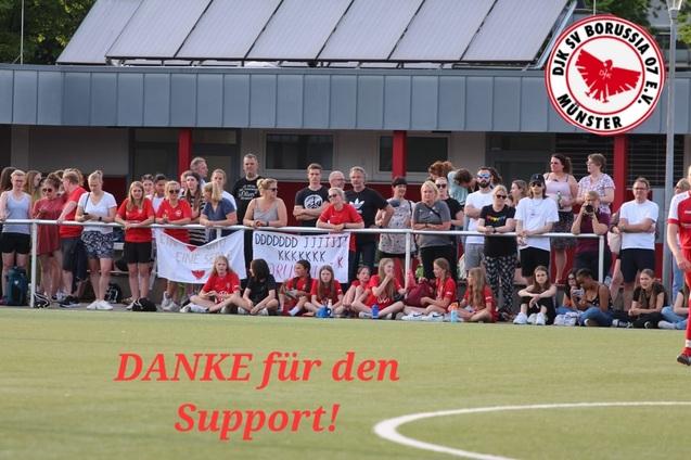 Eine Gruppe von Zuschauern mit roten Trikots steht an einem Fu&szlig;ballfeld, einige halten Banner mit Dankesschriften.