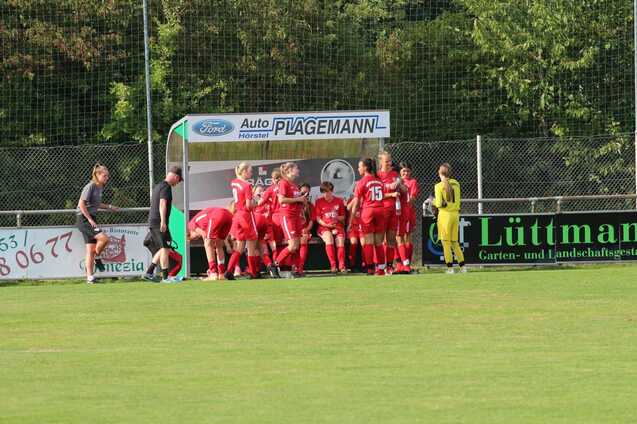Mannschaftsbank mit Spielern in roten Trikots und Trainer auf Fu&szlig;ballplatz, umgeben von Werbebannern.