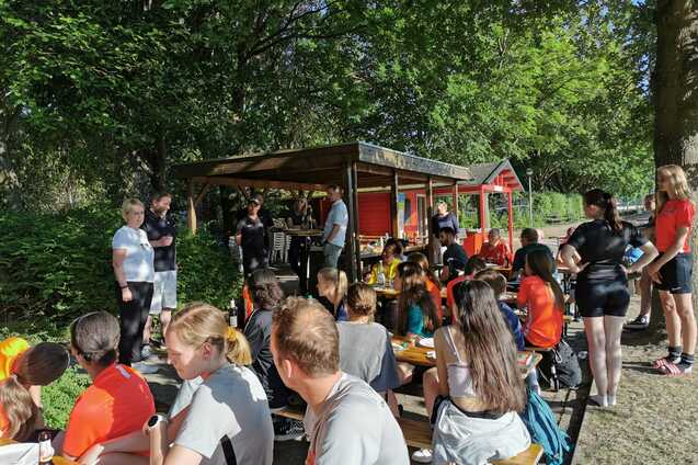 Gruppenszene mit Sportlern, die an Tischen sitzen und essen, umgeben von Gr&uuml;n und einem gem&uuml;tlichen Kiosk.