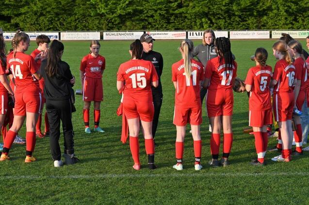 Gruppe von Fu&szlig;ballerinnen in roten Trikots versammelt sich um einen Trainer auf einem gr&uuml;nen Spielfeld.