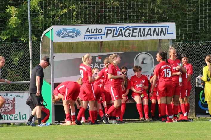 Mannschaftsbank mit Spielern in roten Trikots und Trainer auf Fu&szlig;ballplatz, umgeben von Werbebannern.