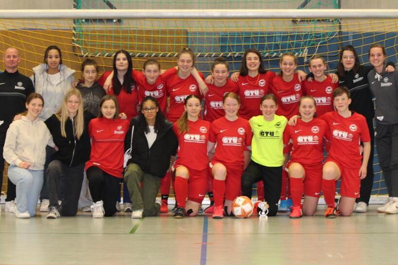 Gruppenfoto einer Fu&szlig;ballmannschaft in roten Trikots, zusammen mit Trainerinnen, in einer Sporthalle vor dem Tor.