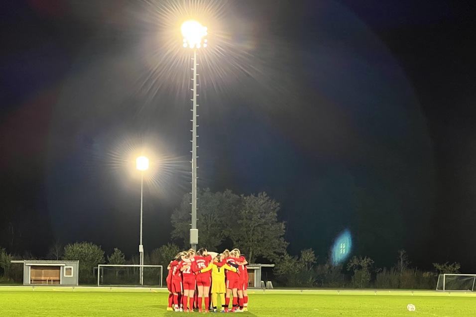 Eine Fu&szlig;ballmannschaft in roten Trikots versammelt sich im n&auml;chtlichen Stadion unter Flutlicht auf einem Rasenfeld.