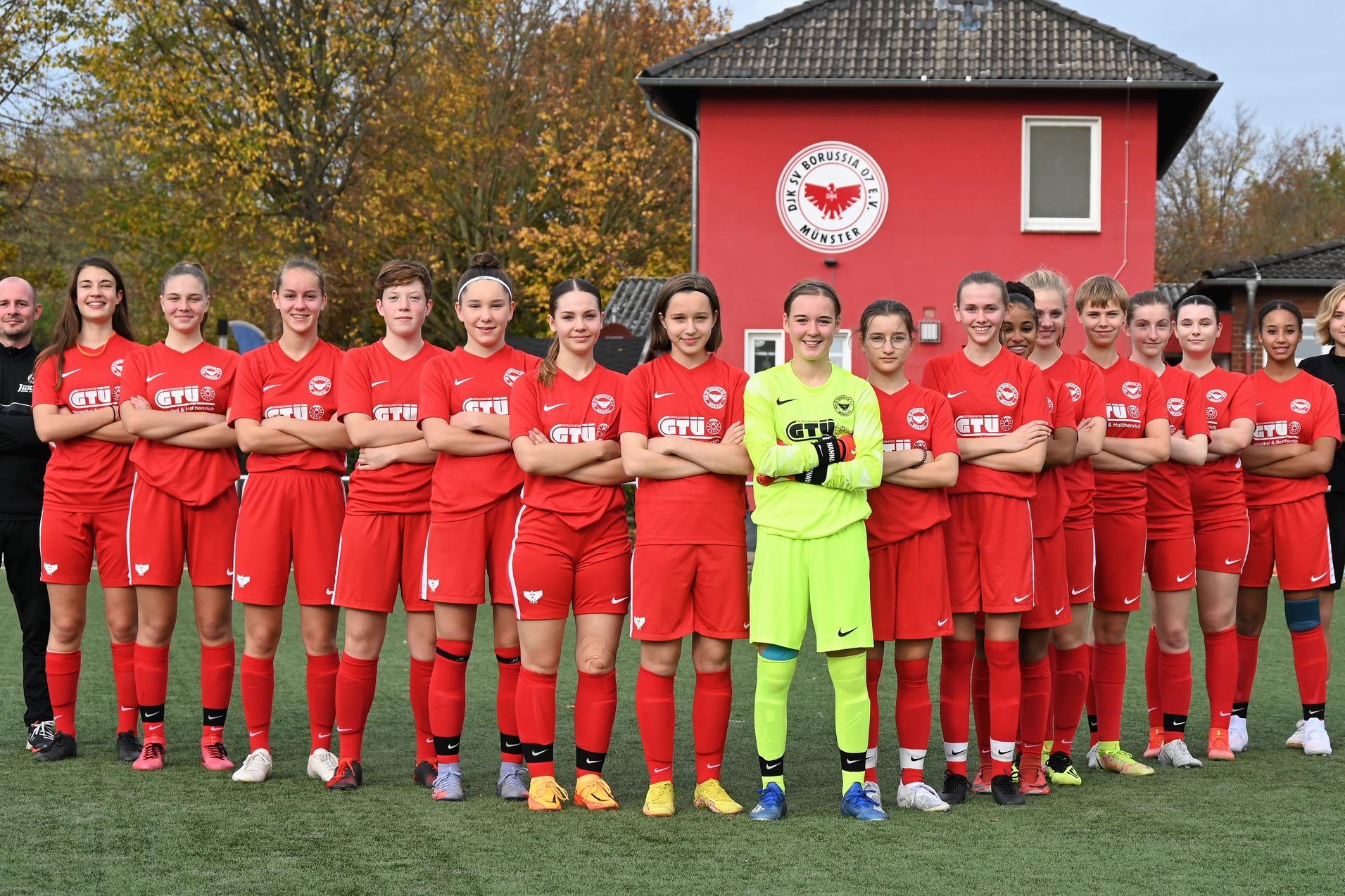 M&auml;dchenfu&szlig;ballmannschaft in roten Trikots steht auf einem Sportplatz, hinter ihnen ein Vereinsgeb&auml;ude mit Logo.