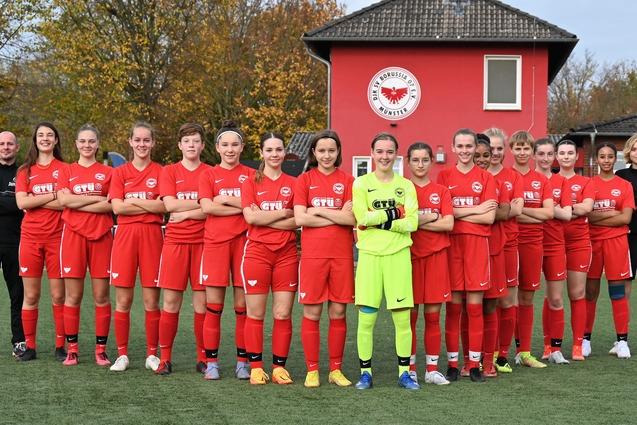 M&auml;dchenfu&szlig;ballmannschaft in roten Trikots steht auf einem Sportplatz, hinter ihnen ein Vereinsgeb&auml;ude mit Logo.