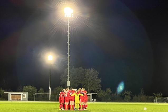 Eine Fu&szlig;ballmannschaft in roten Trikots versammelt sich im n&auml;chtlichen Stadion unter Flutlicht auf einem Rasenfeld.