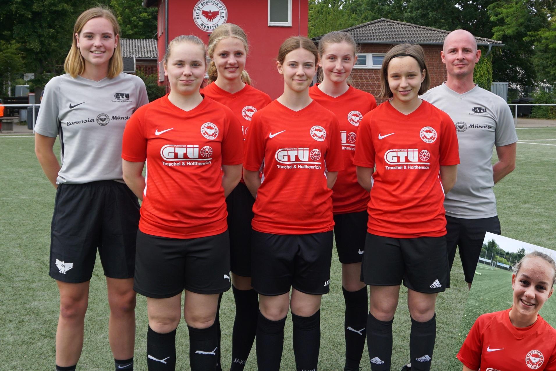 Sechsergruppe von Frauen in roten Fu&szlig;balltrikots, stehend auf einem Sportplatz, mit Trainer im Hintergrund.