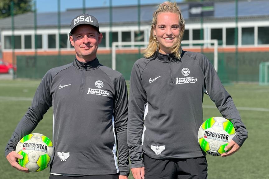Zwei Personen in Sportkleidung halten jeweils einen Fu&szlig;ball auf einem gr&uuml;nen Fu&szlig;ballfeld.