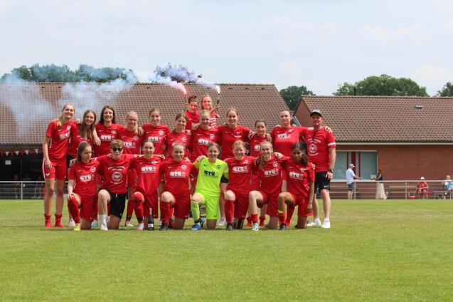 Gruppenfoto einer Fu&szlig;ballmannschaft in roten Trikots, versammelt auf einem Platz mit Rauchfackeln im Hintergrund.