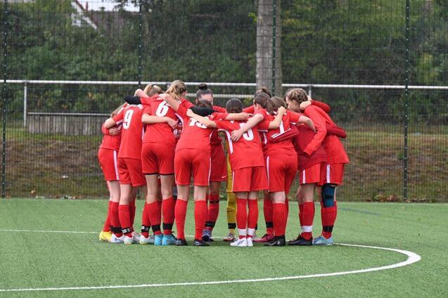 Eine Gruppe von 12 Fu&szlig;ballspielerinnen in roten Trikots steht im Kreis und h&auml;lt sich an den Schultern.