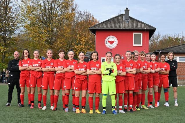 M&auml;dchenfu&szlig;ballmannschaft in roten Trikots steht auf einem Sportplatz, hinter ihnen ein Vereinsgeb&auml;ude mit Logo.