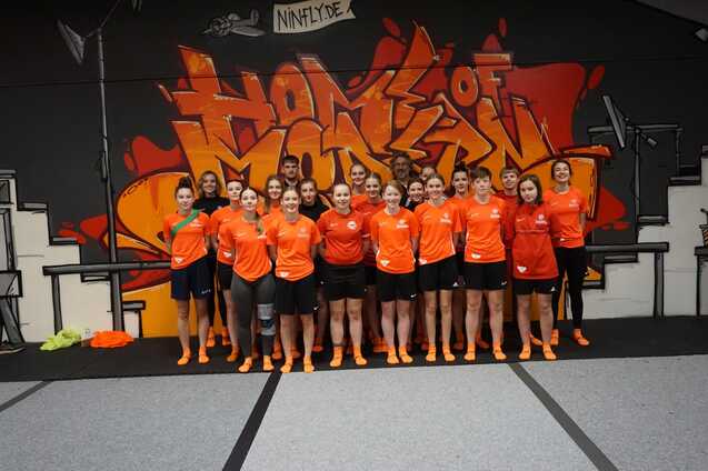 Gruppe von 15 Sportlerinnen in orangefarbenen T-Shirts vor einer graffitiartigen Wand in einer Sporthalle.
