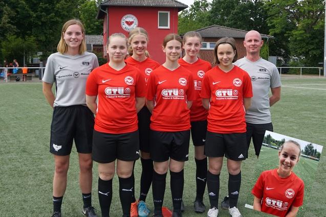 Sechsergruppe von Frauen in roten Fu&szlig;balltrikots, stehend auf einem Sportplatz, mit Trainer im Hintergrund.