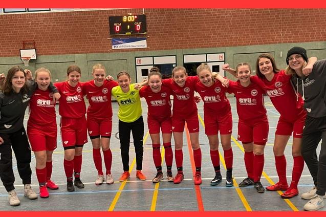 Eine Gruppe von elf Fu&szlig;ballspielerinnen in roten Trikots posiert l&auml;chelnd auf einem Hallenfu&szlig;ballfeld.