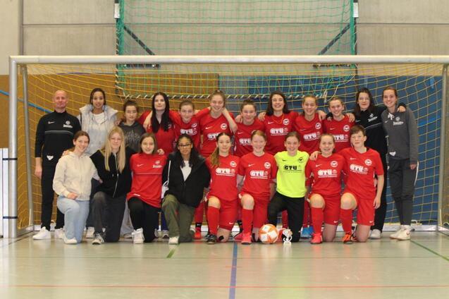 Gruppenfoto einer Fu&szlig;ballmannschaft in roten Trikots, zusammen mit Trainerinnen, in einer Sporthalle vor dem Tor.