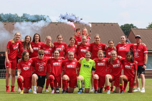 Gruppenfoto einer Fu&szlig;ballmannschaft in roten Trikots, versammelt auf einem Platz mit Rauchfackeln im Hintergrund.