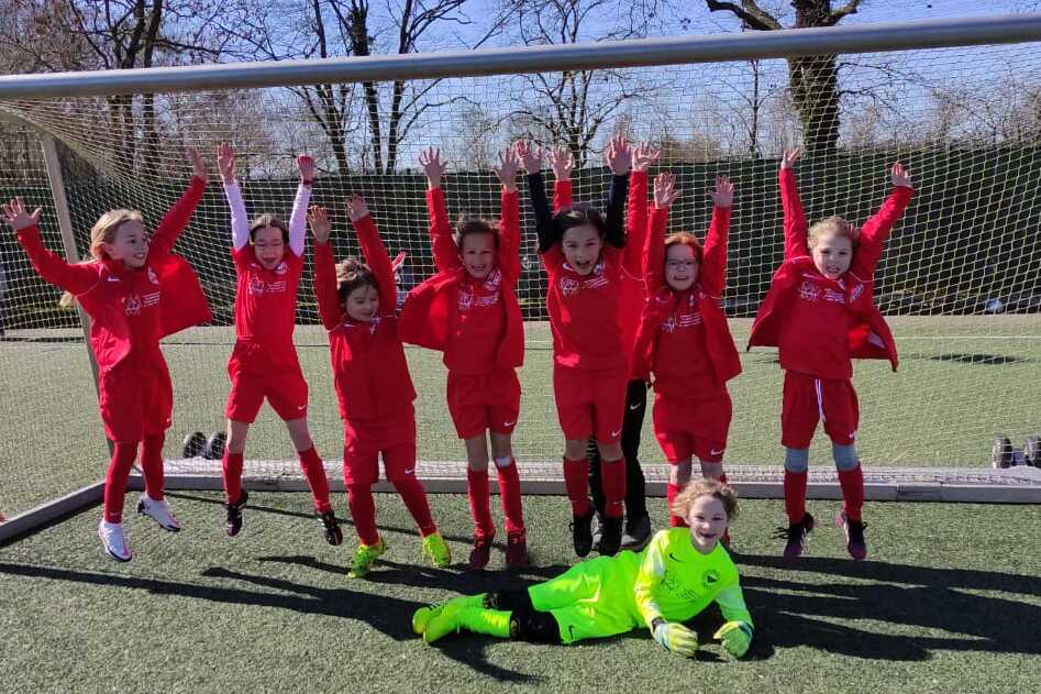 Acht Kinder in roten Fu&szlig;balltrikots stehen jubelnd vor einem Tor auf einem Rasenplatz, ein Torwart in Neon-Gr&uuml;n liegt davor.