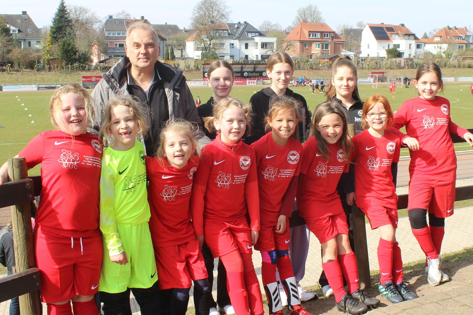 Gruppe von M&auml;dchen in roten Fu&szlig;balltrikots mit Trainer auf einem Sportplatz im Hintergrund.