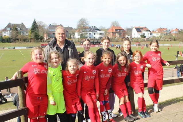 Gruppe von M&auml;dchen in roten Fu&szlig;balltrikots mit Trainer auf einem Sportplatz im Hintergrund.