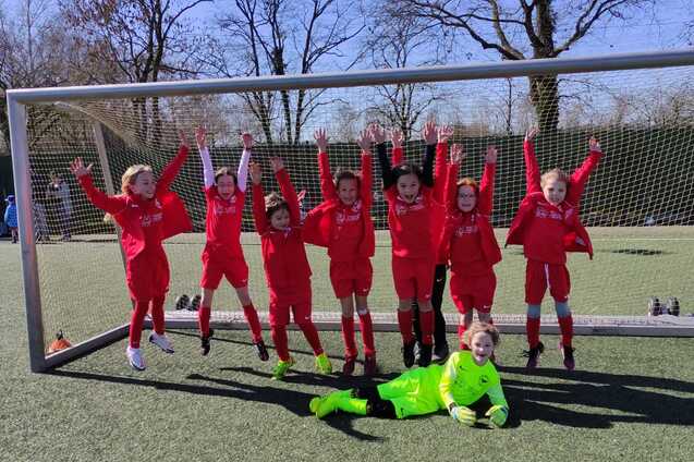 Acht Kinder in roten Fu&szlig;balltrikots stehen jubelnd vor einem Tor auf einem Rasenplatz, ein Torwart in Neon-Gr&uuml;n liegt davor.