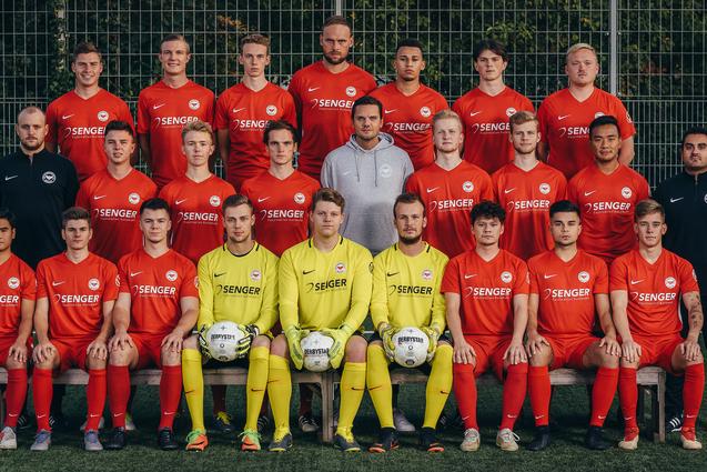 Mannschaftsfoto eines Fu&szlig;ballteams in roten Trikots, posierend auf einem Kunstrasenfeld mit zwei B&auml;llen.