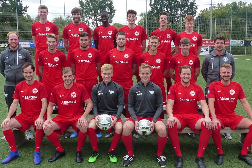 Gruppenfoto einer Fu&szlig;ballmannschaft in roten Trikots, stehend und sitzend auf einem Kunstrasenplatz.