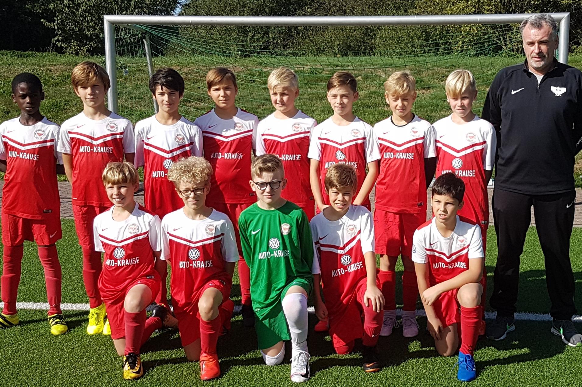 Teamfoto einer Jugendfu&szlig;ballmannschaft in roten Trikots mit Trainer, auf einem Kunstrasenfeld hinter einem Tor.