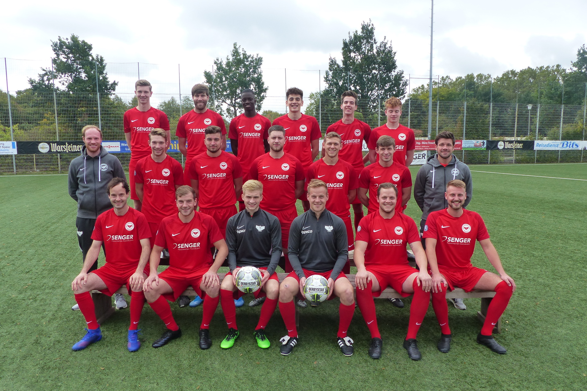 Gruppenfoto einer Fu&szlig;ballmannschaft in roten Trikots, stehend und sitzend auf einem Kunstrasenplatz.