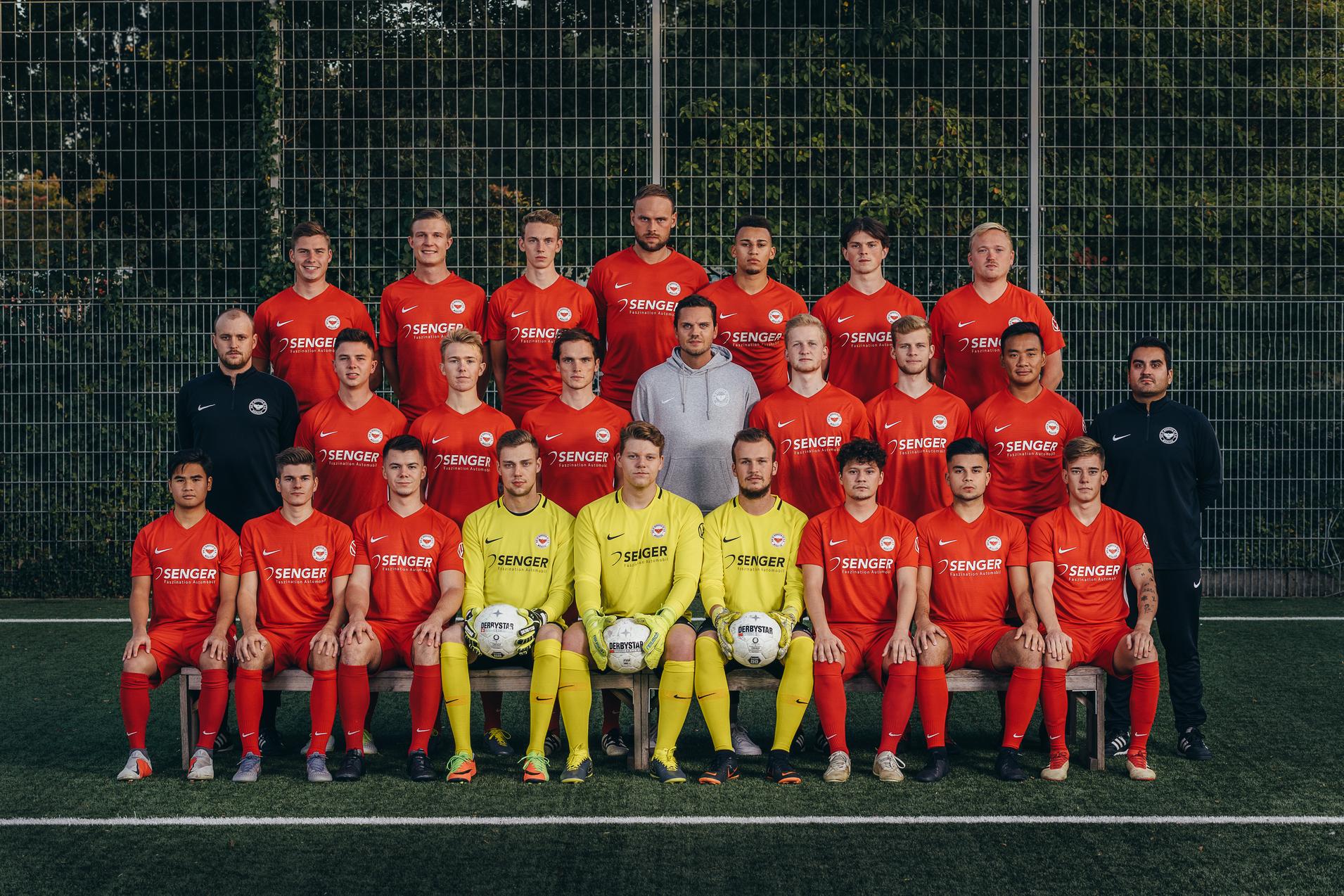 Mannschaftsfoto eines Fu&szlig;ballteams in roten Trikots, posierend auf einem Kunstrasenfeld mit zwei B&auml;llen.