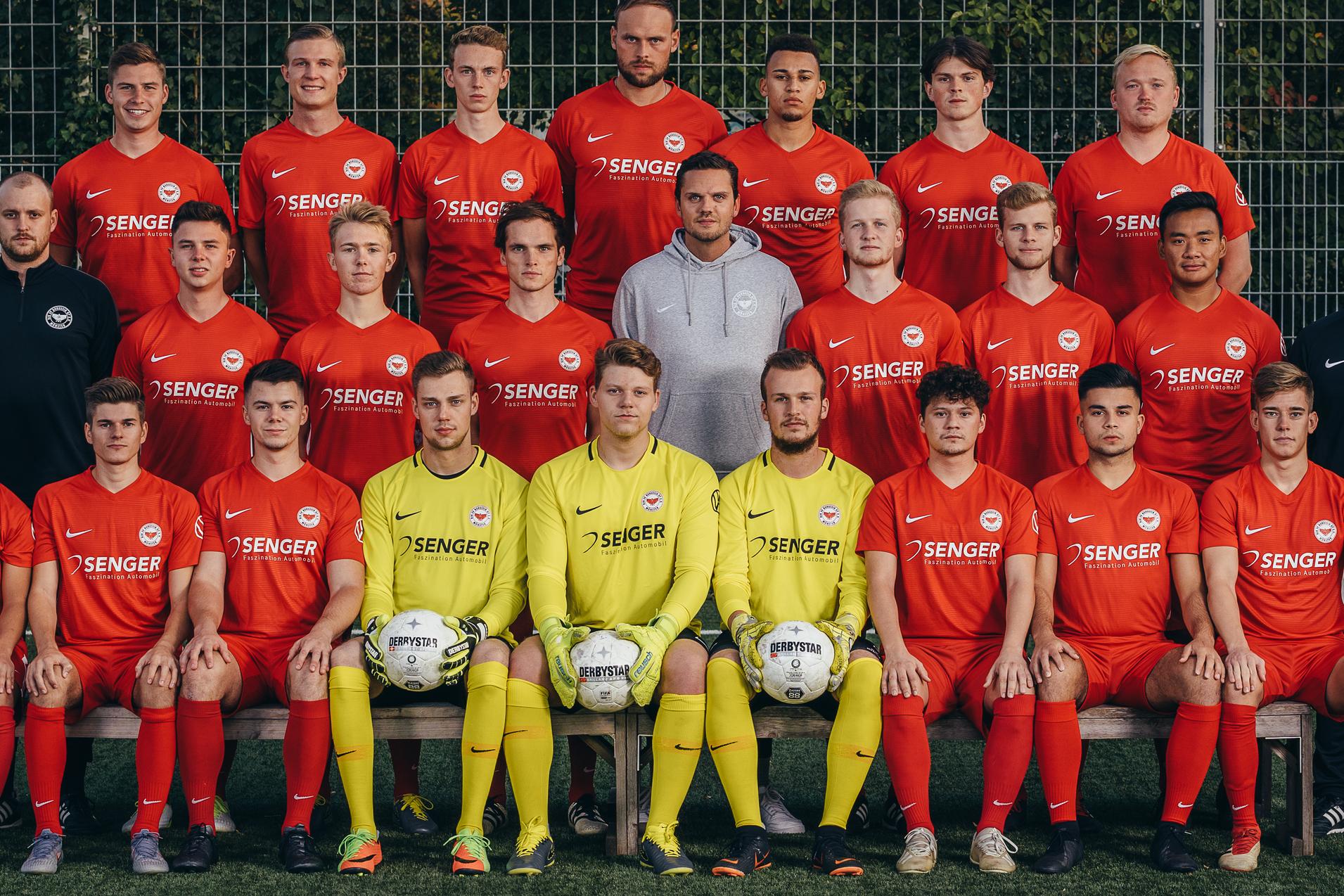 Mannschaftsfoto eines Fu&szlig;ballteams in roten Trikots, posierend auf einem Kunstrasenfeld mit zwei B&auml;llen.