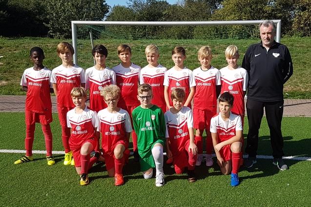 Teamfoto einer Jugendfu&szlig;ballmannschaft in roten Trikots mit Trainer, auf einem Kunstrasenfeld hinter einem Tor.